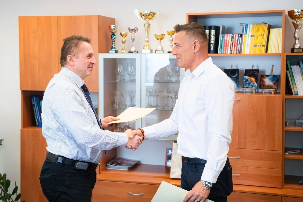 Dvaja muži v kancelárii si podávajú ruky pri podpise dokumentov, za nimi sú vitríny s trofejami.