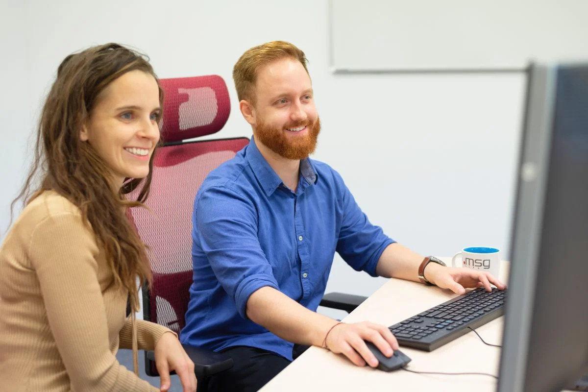 Mladá žena a muž s ryšavou bradou sedia pri jednom počítači a usmievajú sa počas práce.