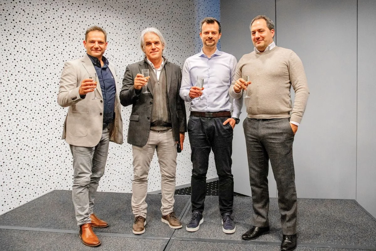 Štyria muži elegantne oblečení stoja spolu a držia šampanské poháre pri firemnom podujatí.